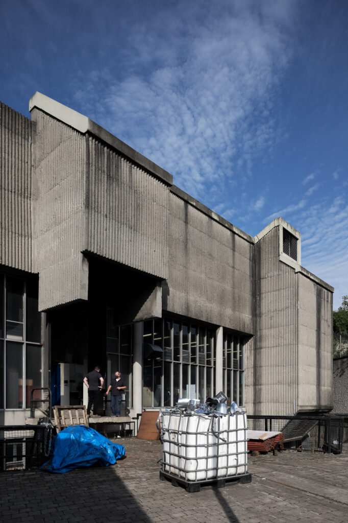 linnenhall-vakschool-golfbeton-gebouw-met-twee-mannen-in-zwart-bij-de-ingang-en-witte-opslagcontainer-vóór