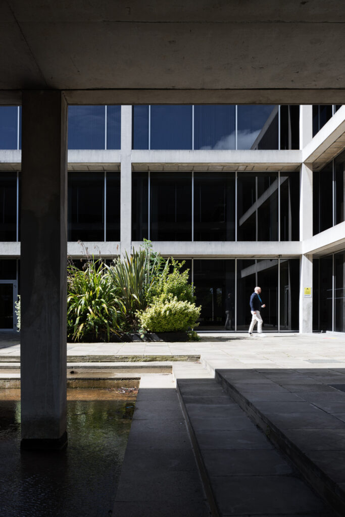 man-walks-through-sunlit-courtyard-with-plants-surrounded-by-exposed-concrete-and-glass-modernist-building