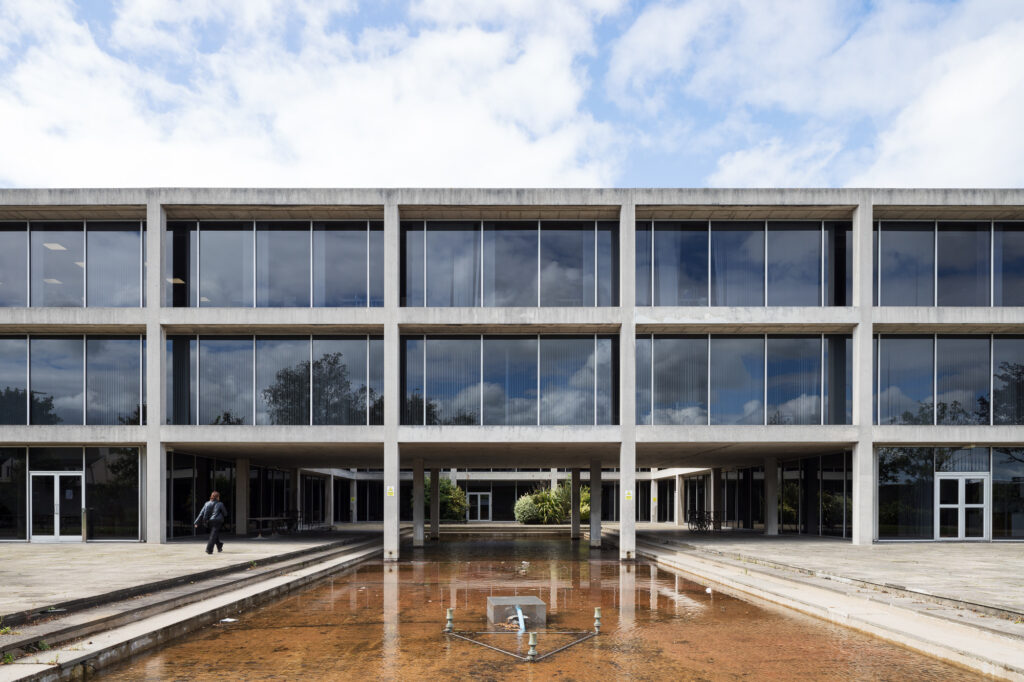 woman-walking-across-courtyard-towards-merrion-hall-front-elevation-of-exposed-concrete-and-reflective-glass-with-central-fountain
