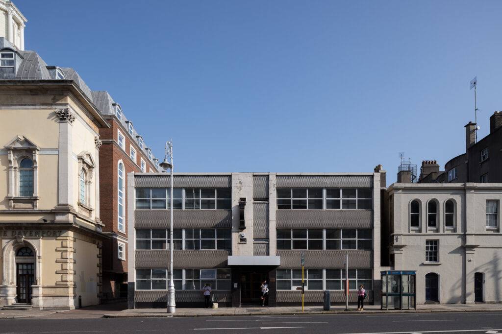 elevation-of-the-three-storey-morrissey's-blue-grey-and-off-white-mosaic-tiled-facade-between-two-buildings-in-front-of-a-bus-stop