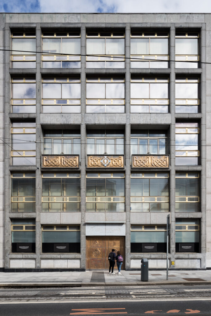 elevation-of-five-storey-new-ireland-assurance-building-with-limestone-panels-and-golden-framed-windows-as-people-stand-looking-at-golden-door