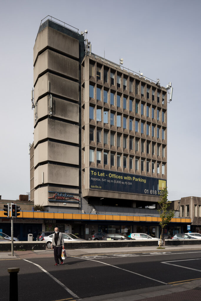 eight-storey-concrete-phibsboro-centre-with-nine-storey-vertical-circulation-tower-as-man-with-shopping-bag-crosses-the-road