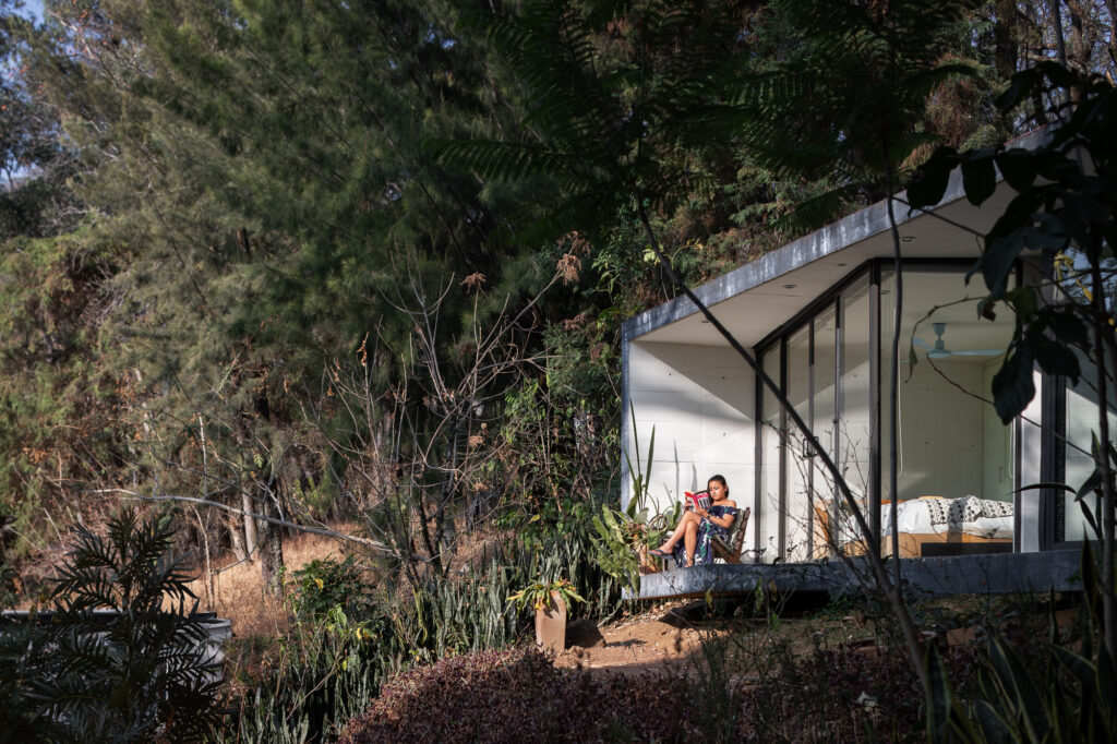 woman-in-blue-dress-reading-in-front-of-black-and-white-concrete-and-glass-bungalow-in-the-forest