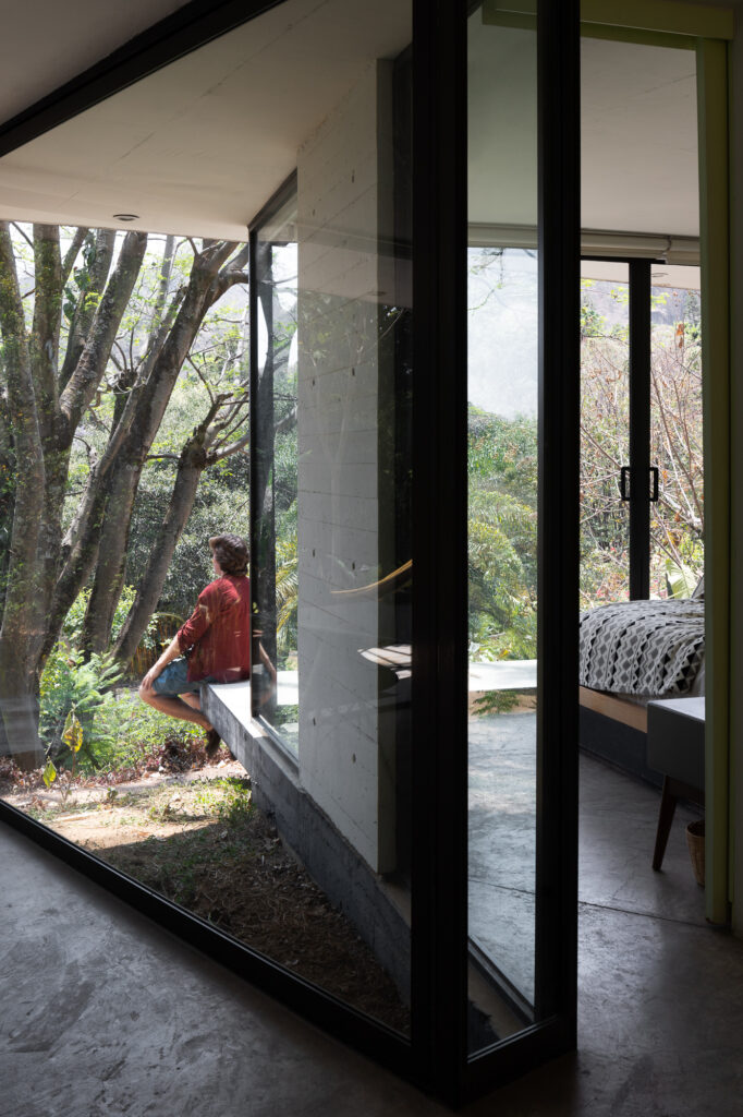 view-from-interior-through-floor-to-ceiling-glazing-of-man-sitting-on-cantilevering-concrete-plinth-overlooking-forest