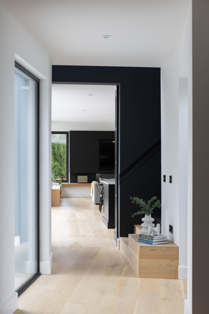 hallway-with-wooden-steps-with-books-and-white-sculpture-on-top-and sculptured-vase with-plant-beside-black-stairway-with-living-area-in-background