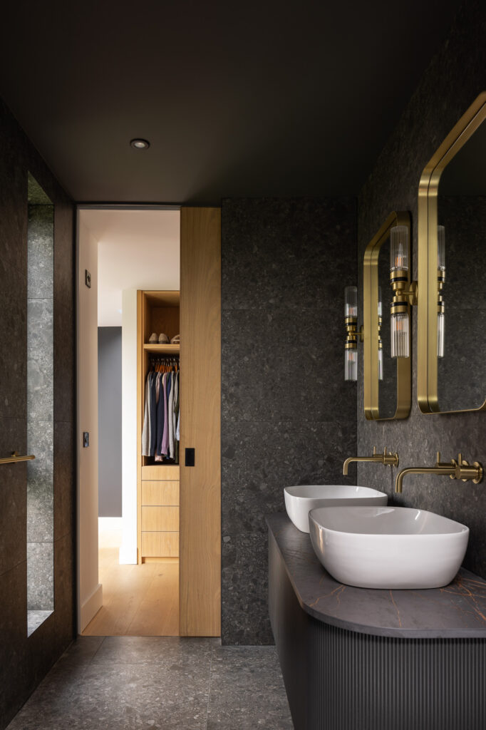 black-tiled-bathroom-golden-taps-and-mirror-frames-with-open-wooden-sliding-revealing-walk-in-closet