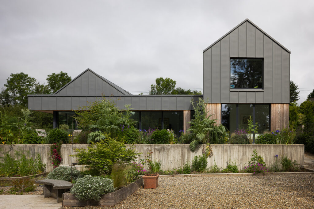 residence-with-two-pitched-roof-sections-with-timber-and-zinc-cladding-perched-on-concrete-plinth-above-plant-filled-garden