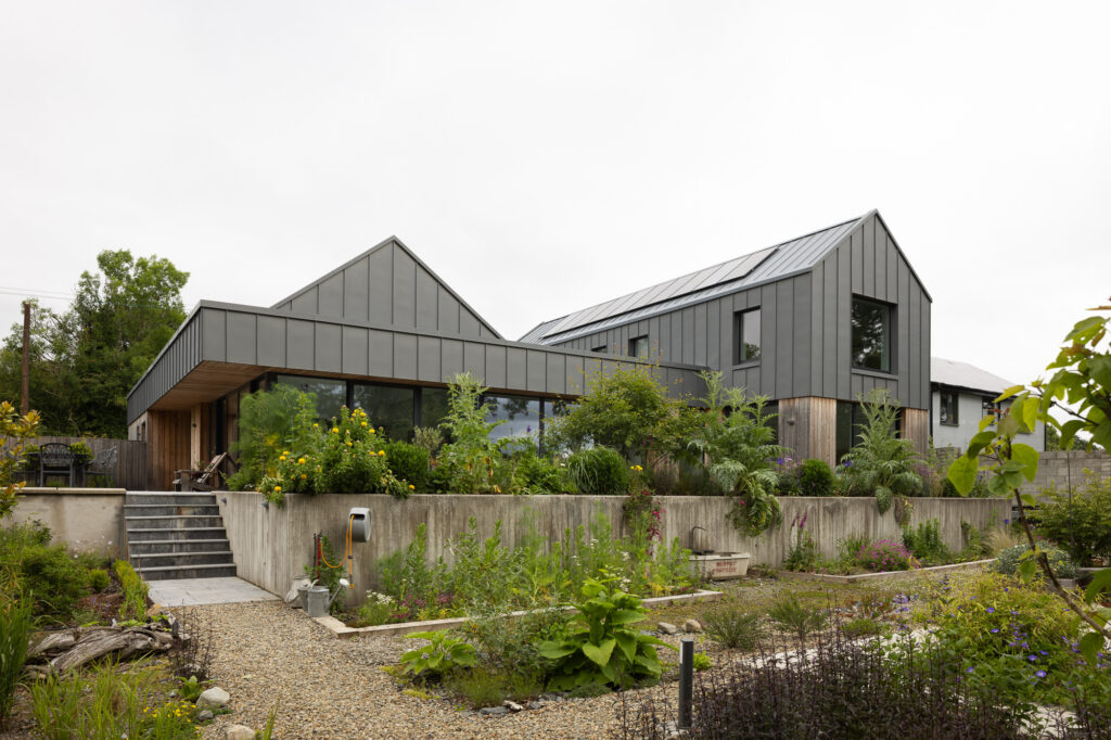 residence-with-two-pitched-roof-sections-with-timber-and-zinc-cladding-perched-on-concrete-plinth-above-plant-filled-garden